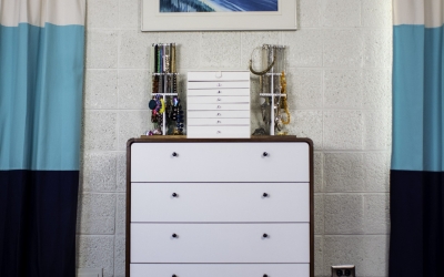 A symmetrical bedroom with a brown dresser with white drawers, a white wall with a painting hanging above the dresser, and curtains that are white, blue and black.
